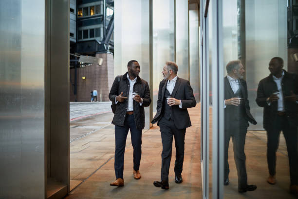 Male Business Colleagues Walking and Talking with Coffee African and Caucasian businessmen in 40s and 50s walking side by side and discussing ideas outdoors on coffee break. colleagues outside stock pictures, royalty-free photos & images
