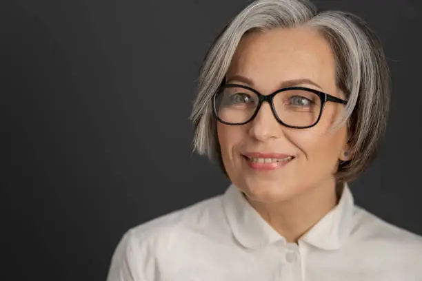 Pretty gray-haired woman in white shirt. intelligent middle-aged businesswoman in glasses with medium length care hairstyle smiling on grey background. Close up portrait. Textspace at left side.