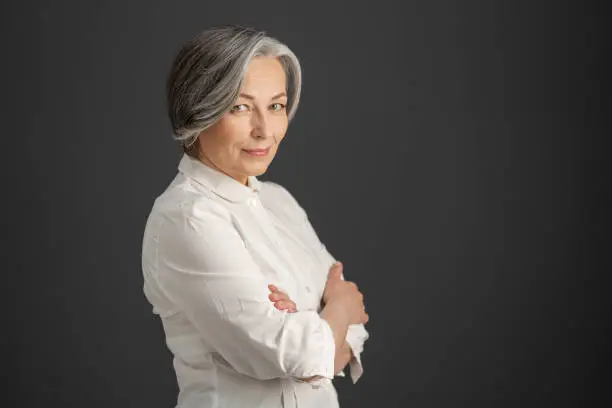 Pretty white-haired woman smiles looking at camera with arms crossed. Stylish caucasian businesswoman isolated on dark gray background. Textspace at right side. Business concept.