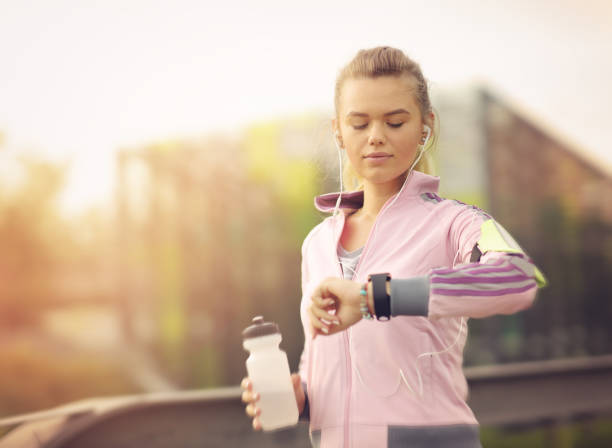 Female runner running at sunset in city park - Healthy fitness woman jogging outdoors - Athlete listening to music during workout at park and adjusting smart watch stock photo
