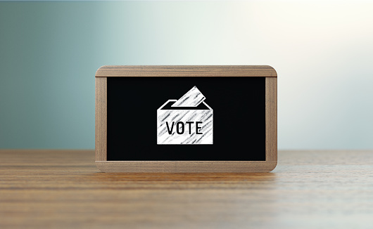 Wooden framed blackboard with voting icon sitting over wooden surface in front of defocused background. Horizontal composition with copy space. Election and voting concept.