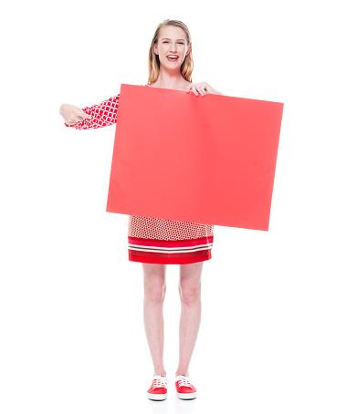 Front view of aged 18-19 years old who is beautiful caucasian young women standing in front of white background wearing dress who is happy who is showing with hand and holding sign with copy space