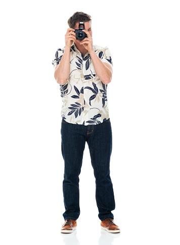 Full length of aged 30-39 years old with brown hair caucasian young male tourist standing in front of white background wearing jeans who is laughing who is photographing and holding camera
