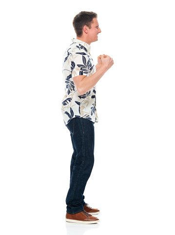 Side view of aged 30-39 years old with brown hair caucasian young male standing in front of white background wearing jeans who is excited and cheering and showing fist who is and doing fist pump