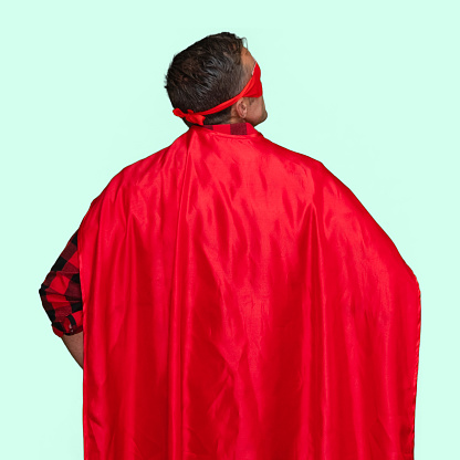 Rear view of with short hair caucasian male hero standing in front of colored background wearing mask - disguise who is conquering adversity with hand on hip