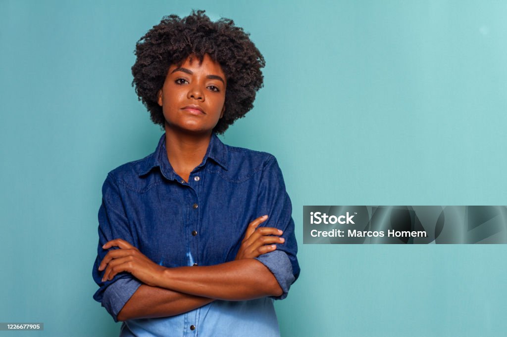 Black young woman with black power hair wearing a blue jeans shirt on blue background Black woman with curly hair wearing a blue jeans shirt on blue background Women Stock Photo