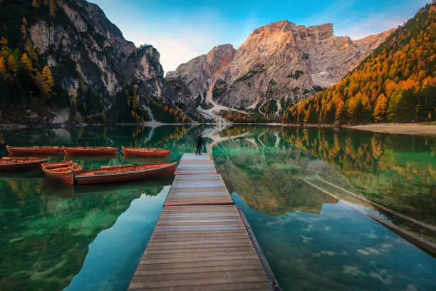 Traveler photographer taking a photos and enjoying the view on the pier of lake Braies. Stunning place with colorful larches, mountains and wooden boats on the lake, Dolomites, Italy, Europe