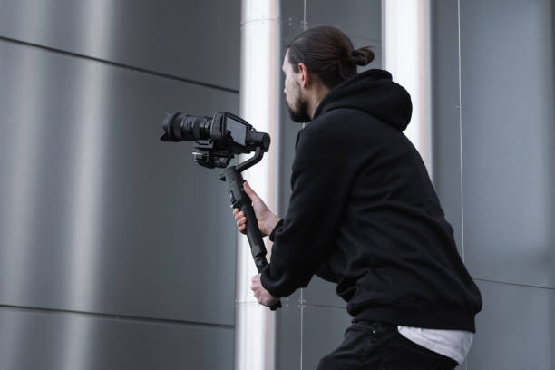 istock Young Professional videographer holding professional camera on 3-axis gimbal stabilizer. Pro equipment helps to make high quality video without shaking. Cameraman wearing white hoodie making a videos. 1226716584
