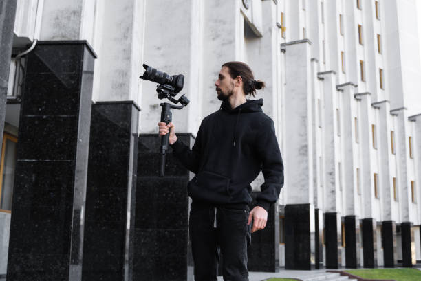 istock Young Professional videographer holding professional camera on 3-axis gimbal stabilizer. Pro equipment helps to make high quality video without shaking. Cameraman wearing black hoodie making a videos. 1226717058