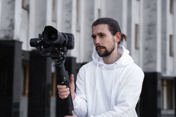 istock Young Professional videographer holding professional camera on 3-axis gimbal stabilizer. Pro equipment helps to make high quality video without shaking. Cameraman wearing white hoodie making a videos. 1226718279