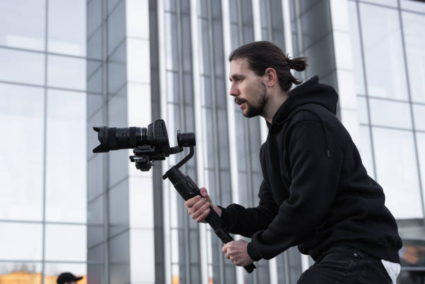 istock Young Professional videographer holding professional camera on 3-axis gimbal stabilizer. Pro equipment helps to make high quality video without shaking. Cameraman wearing white hoodie making a videos. 1226719283