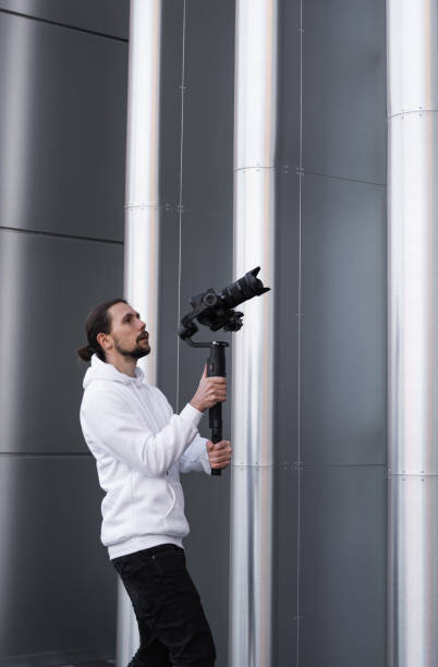 istock Young Professional videographer holding professional camera on 3-axis gimbal stabilizer. Pro equipment helps to make high quality video without shaking. Cameraman wearing white hoodie making a videos. 1226719903