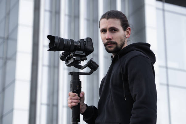 istock Young Professional videographer holding professional camera on 3-axis gimbal stabilizer. Pro equipment helps to make high quality video without shaking. Cameraman wearing white hoodie making a videos. 1226720268