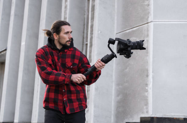 istock Young Professional videographer holding professional camera on 3-axis gimbal stabilizer. Pro equipment helps to make high quality video without shaking. Cameraman wearing red shirt making a videos. 1226720967
