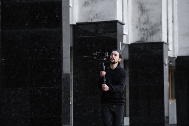 istock Young Professional videographer holding professional camera on 3-axis gimbal stabilizer. Pro equipment helps to make high quality video without shaking. Cameraman wearing black hoodie making a videos. 1226721105
