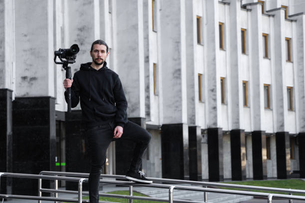 istock Young Professional videographer holding professional camera on 3-axis gimbal stabilizer. Pro equipment helps to make high quality video without shaking. Cameraman wearing black hoodie making a videos. 1226721177