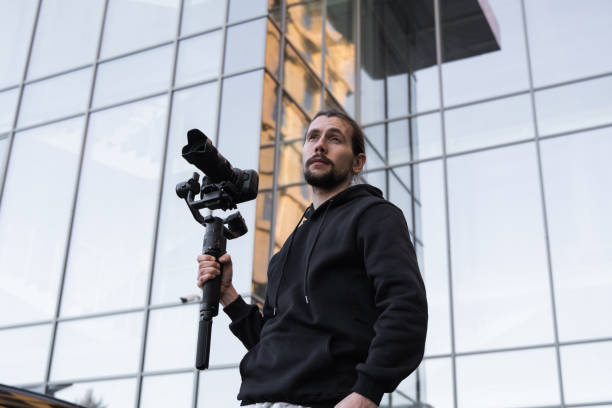 istock Young Professional videographer holding professional camera on 3-axis gimbal stabilizer. Pro equipment helps to make high quality video without shaking. Cameraman wearing white hoodie making a videos. 1226721238