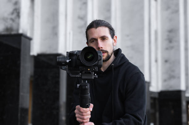 istock Young Professional videographer holding professional camera on 3-axis gimbal stabilizer. Pro equipment helps to make high quality video without shaking. Cameraman wearing black hoodie making a videos. 1226723223