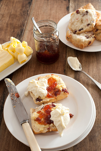 Fresh buttered rock cake with a dollop of whipped cream and fruit jam served on a white plate with a pat of butter and jar of jelly behind