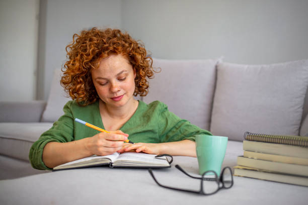 Happy cute lady at home write notes on a diary while drink a cup of tea and rest and relax taking a break. Red curly beautiful lady sit down in the house. People enjoying home lifestyle writing and study