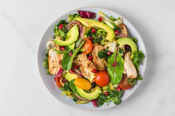 Photo of Salad with grilled chicken breast, avocado, pomegranate seeds and tomato on white background. Top view