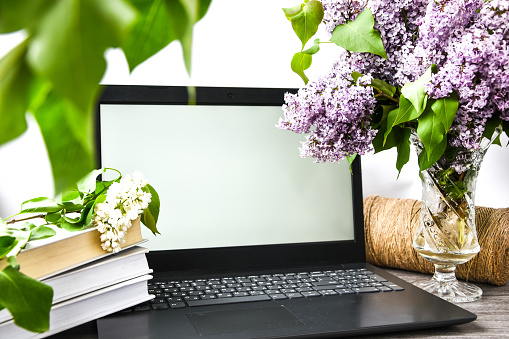 Lilac flower bouquet in glass vase with open laptop white screen and books, rope, copy space for text, springtime, blossom