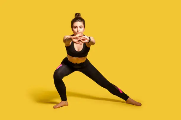 Athletic pretty girl with hair bun in tight sportswear doing sport, lower-body exercise, stretching hands out, warming up training muscles for flexibility. full length studio shot, isolated on yellow