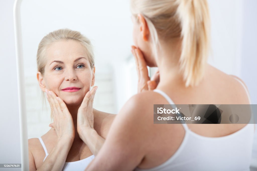 Midden oude vrouw die rimpels in spiegel bekijkt. Plastische chirurgie en collageen injecties. Make-up. Macrogezicht. Selectieve focus - Royalty-free Anti-veroudering Stockfoto