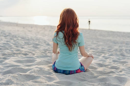 Young woman relaxing on the sand on a beach sitting looking out over the ocean with her back to the camera