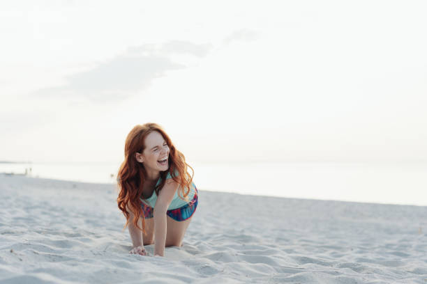 Laughing young woman crawling across beach sand with the ocean at her rear looking to the side with a beaming smile