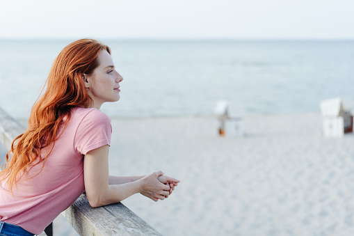Young redhead woman enjoying a day at the beach standing on a deck leaning on a railing overlooking the sand