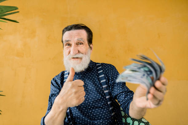 joyful satisfied handsome senior man with well-groomed beard, wearing trendy clothes, holding a lot of paper money, dollars bills, and showing his thumb up. studio shot on yellow background - senior getting groomed studio imagens e fotografias de stock