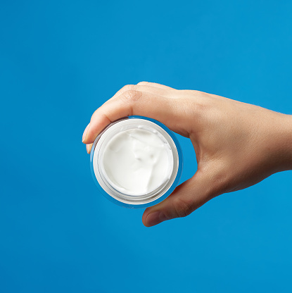 female hand holds  a round jar with white cream for face and body on a blue background, concept of moisturizing