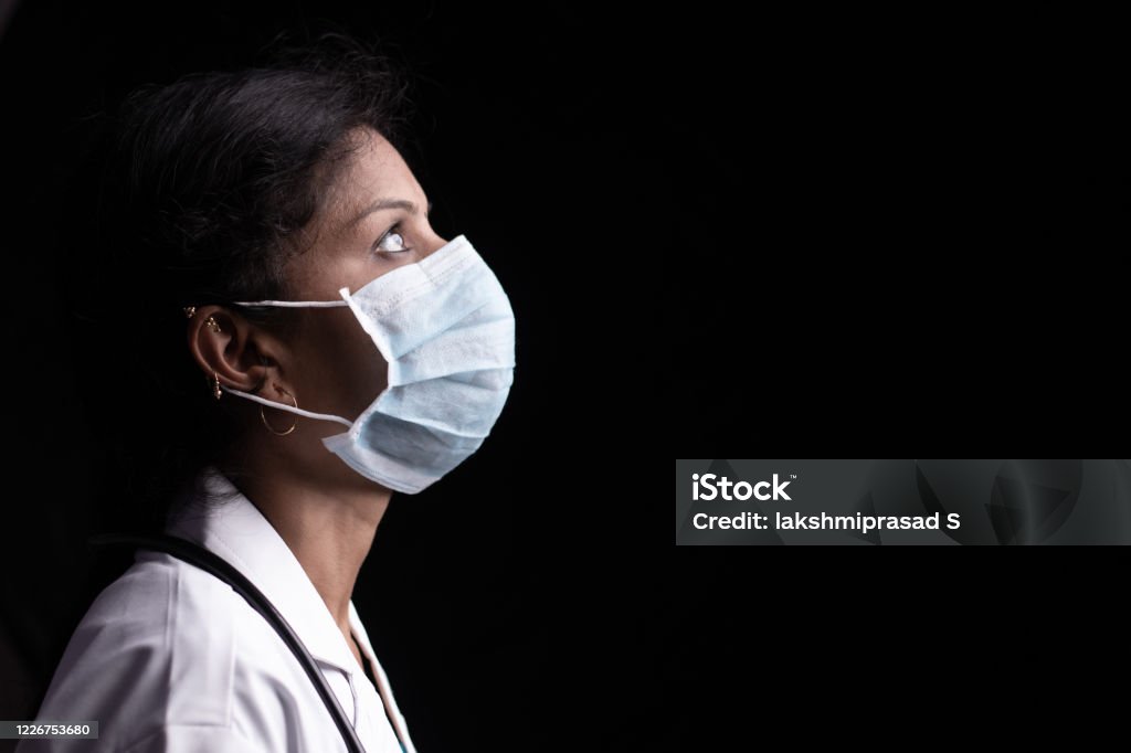 Profile view of young woman doctor with opened eyes in medical mask on black background looking up - concept of hope and fight to end coronavirus or covid-19 crisis. Profile view of young woman doctor with opened eyes in medical mask on black background looking up - concept of hope and fight to end coronavirus or covid-19 crisis Doctor Stock Photo