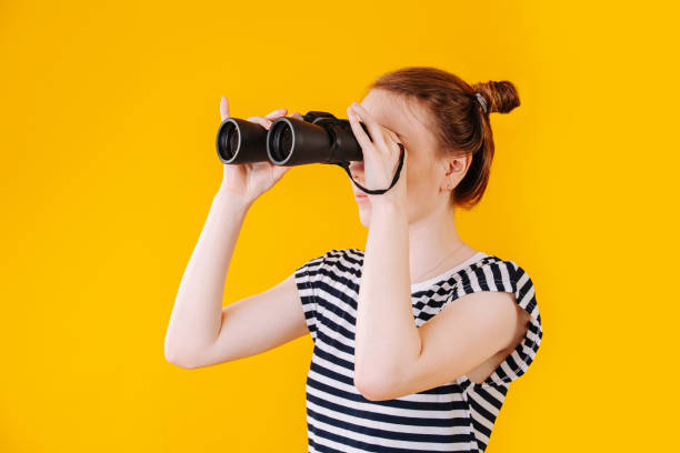 Girl in search with binoculars in hand Girl in search with binoculars in hand, studio portrait on yellow background searching binoculars stock pictures, royalty-free photos & images