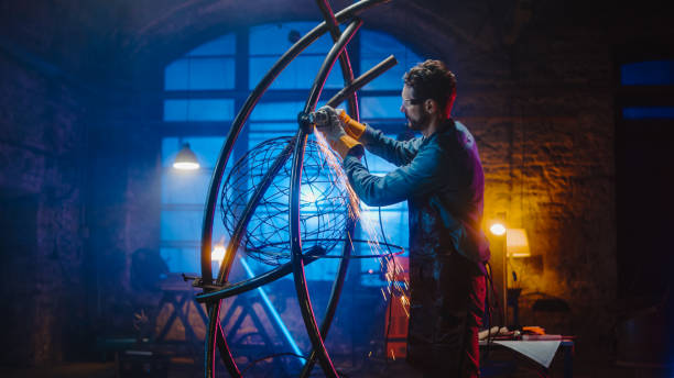 handsome male artist uses an angle grinder to make brutal metal sculpture in studio. hipster guy polishes metal tube with sparks flying off it. contemporary fabricator creating abstract steel art. - sculpture imagens e fotografias de stock