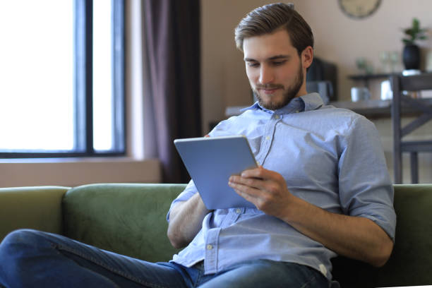 istock Smiling handsome man holding and using tablet with social networks at home, happy freelancer making online shopping, sitting on cozy sofa at living room 1226764788