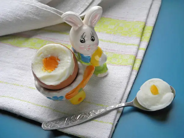 Broken hard boiled egg in decorative eggcup in form of rabbit with wheelbarrow, spoon with piece of egg on linen napkin. Egg white and yolk. Baby and diet food.