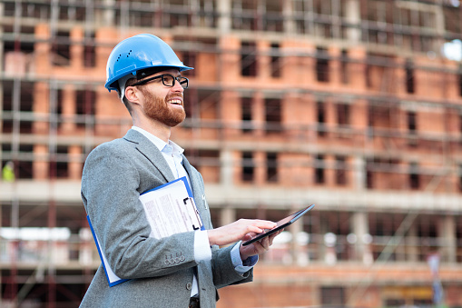 Smiling happy young Caucasian architect or businessman visiting construction site, working on a tablet and holding clipboard with documents under his arm