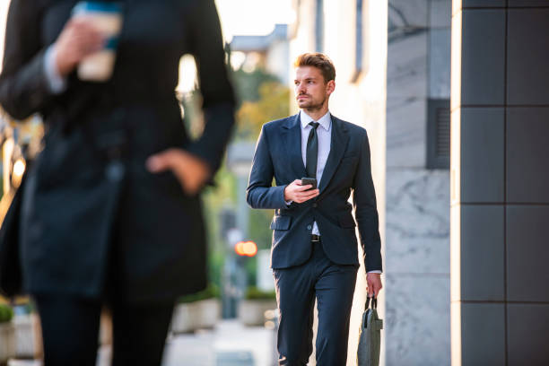 Low angle focus on background of serious male professional in suit and tie holding smart phone while walking and looking away from camera.