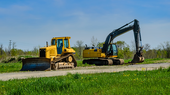 Two pieces of heavy equipment in a field next to a gravel road on a sunny day.