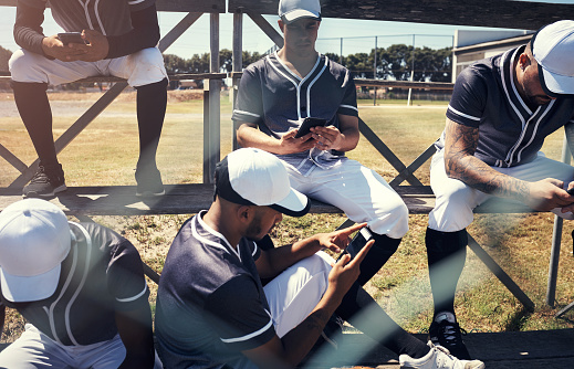 Shot of a young man using a smartphone at a baseball game surrounded by his team mates