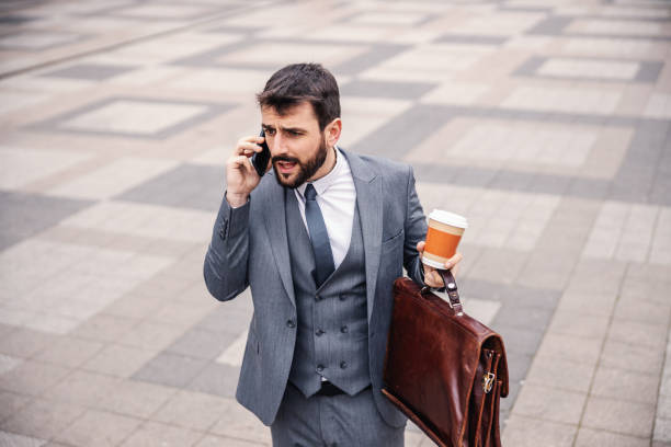 istock Young serious bearded businessman holding bag and his morning coffee while going to work and having phone conversation. 1226783465
