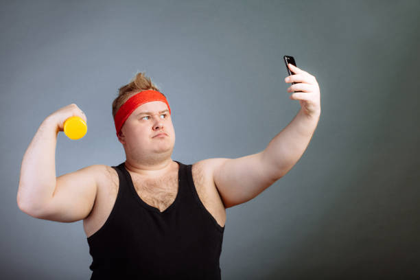 istock fat man with big belly, holding dumbbell, doing selfie on grey background 1226783623