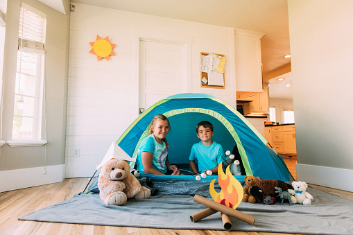 A young brother and sister are playing in a tent set up in their home during the day. Due to the coronavirus pandemic, they are in need of staying home and staying safe. They are having fun camping and roasting marshmallows inside the home with a staycation.