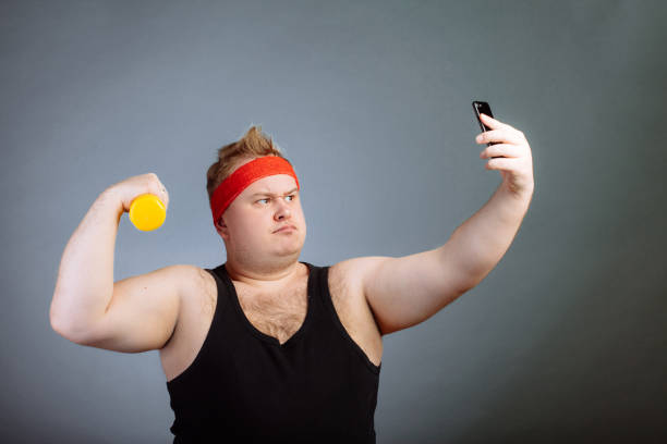istock fat man with big belly, holding dumbbell, doing selfie on grey background 1226783860