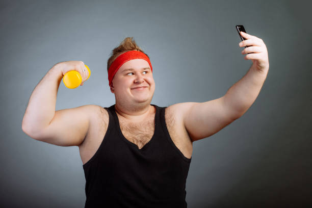 istock fat man with big belly, holding dumbbell, doing selfie on grey background 1226784397
