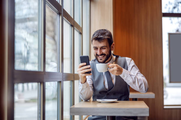 istock Young handsome bearded CEO sitting on coffee break, holding cup and reading messages from important client. 1226784415