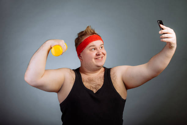 istock fat man with big belly, holding dumbbell, doing selfie on grey background 1226784486