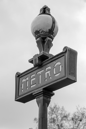 The Paris Metro sign with a a street lamp, indicating the presence of the subway system entrance.  Black and white.